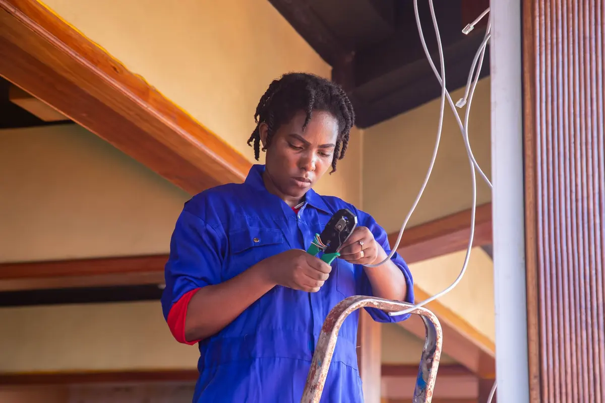 Electrician in blue coveralls stripping a wire while standing on a ladder
